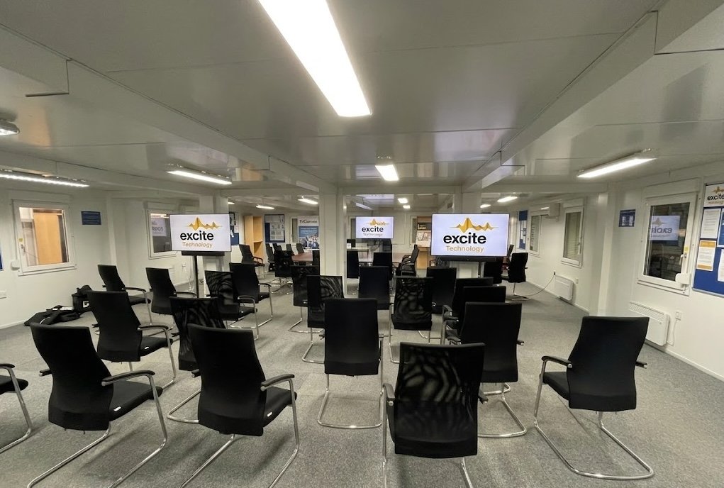 Rows of empty black chairs face multiple screens displaying the Excite Technology logo in a bright, modern conference room with white walls and windows.