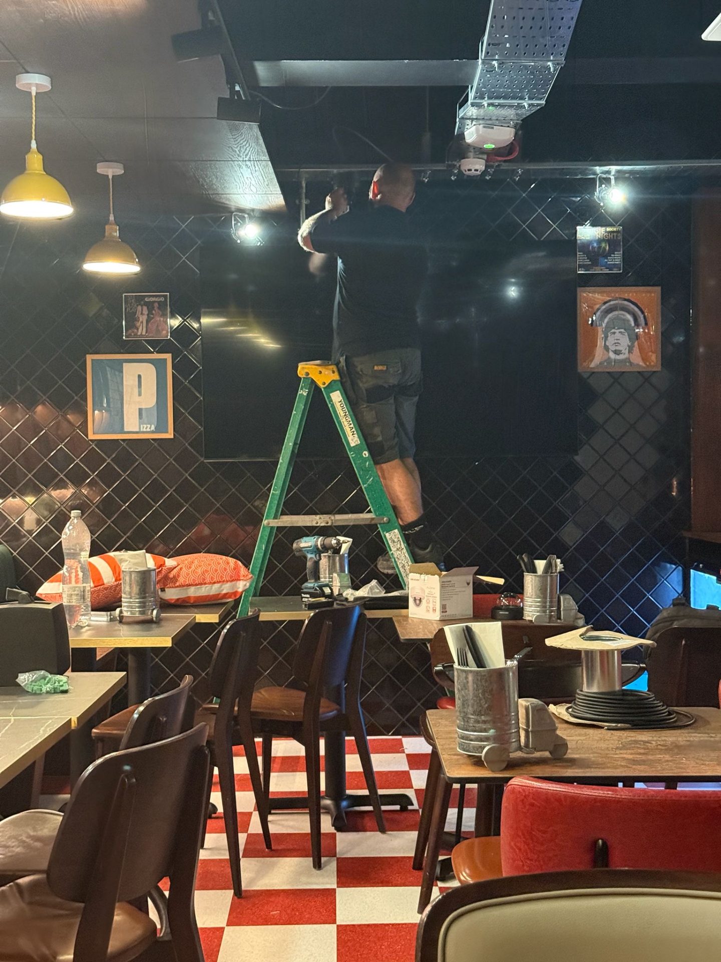 A person stands on a green ladder fixing the ceiling in a Pizza Pilgrims restaurant with black tiled walls, chequered red-and-white floor, and scattered tables with tools and paint tins. Framed pictures hang on the wall.