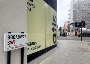 Street corner with a Broadway SW1 sign, construction barriers, traffic lights, cement lorry, and scaffolding on a building. Large yellow sign reads, Opening Spring 2026. Set in an urban setting near St James’s Park in London’s City of Westminster, this location is currently under development by ATIS.