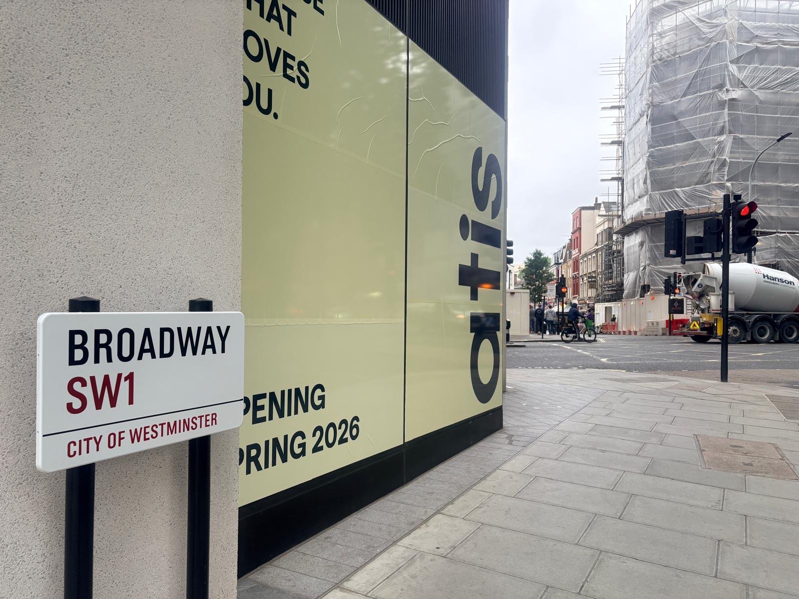 Street corner with a Broadway SW1 sign, construction barriers, traffic lights, cement lorry, and scaffolding on a building. Large yellow sign reads, Opening Spring 2026. Set in an urban setting near St James’s Park in London’s City of Westminster, this location is currently under development by ATIS.