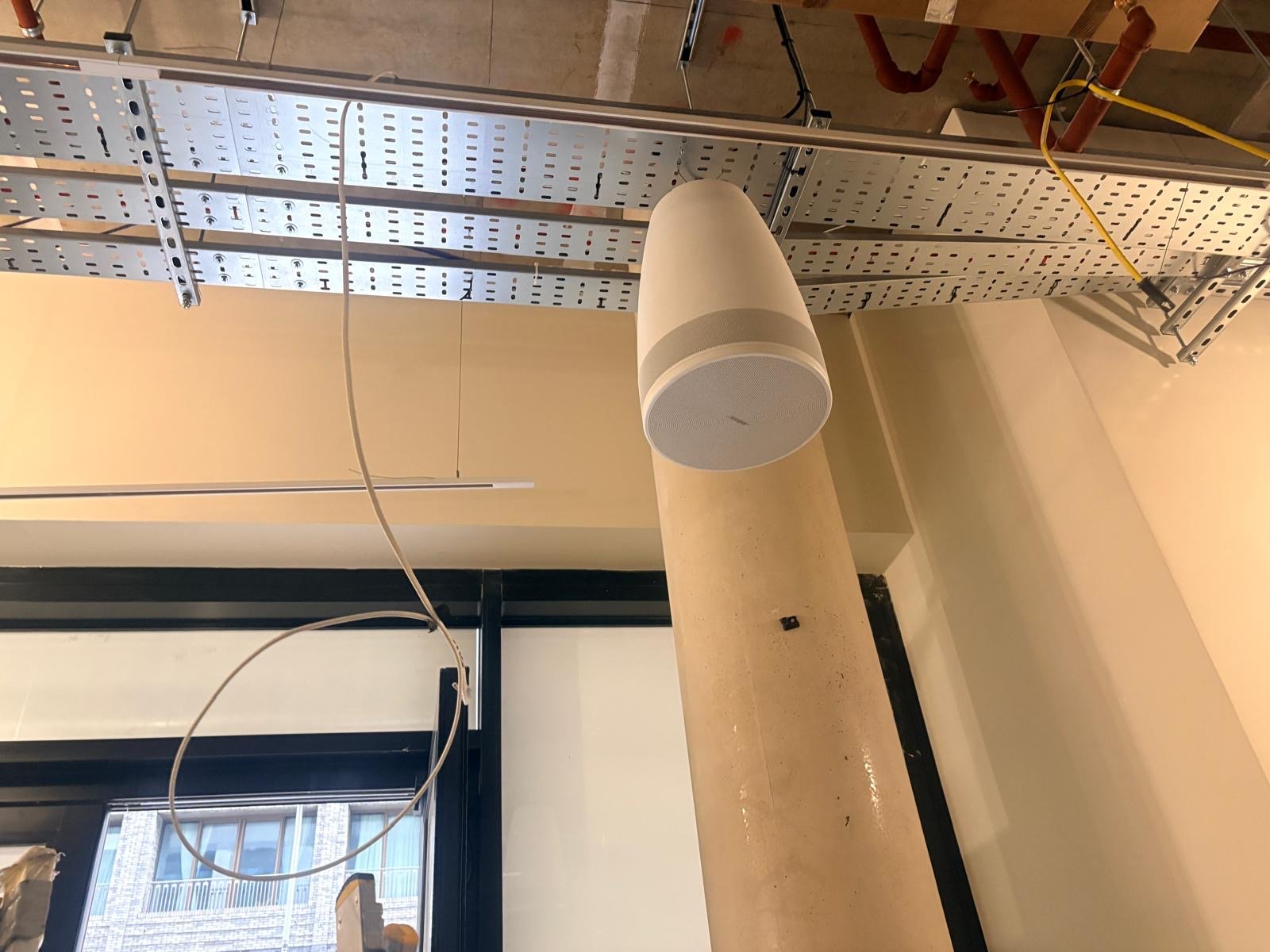 A white cylindrical speaker or device is mounted on a beige pillar under a metal cable tray, in an industrial-style room with exposed pipes and large windows, reminiscent of the modern workspaces found around St James’s Park in London—perfect for broadcasting ATIS updates throughout the area.