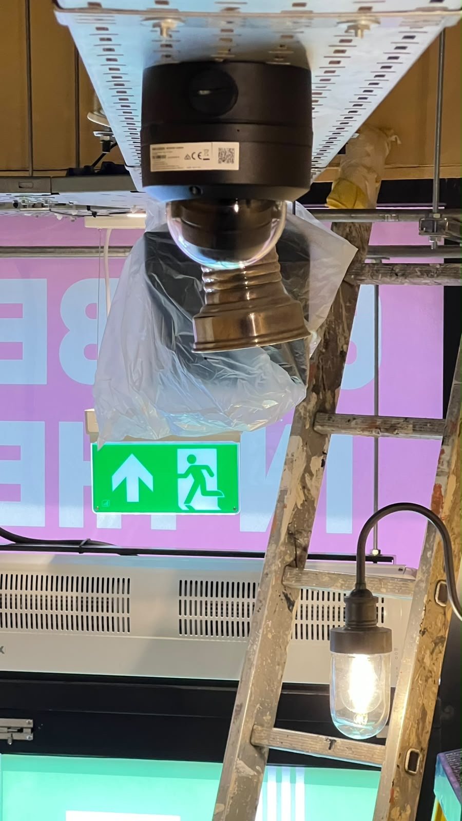 An upside-down view of a metal ladder, a hanging light bulb, a CCTV camera wrapped in plastic, and a green emergency exit sign with a running figure, set against a pink background—evoking the quirky energy of Wimbledon or the playful vibe found at Pizza Pilgrims.