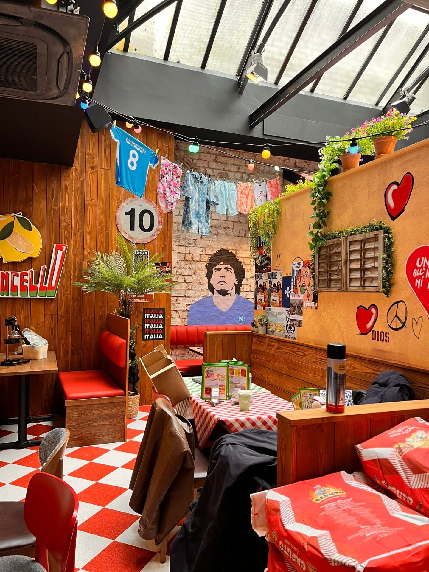 An Italian-themed restaurant in Glasgow with red chequered tablecloths, football decor, hanging laundry, and a mural of Diego Maradona on a brick wall. Fairy lights and plants add a cosy atmosphere that’s perfect for Pizza Pilgrims enthusiasts.