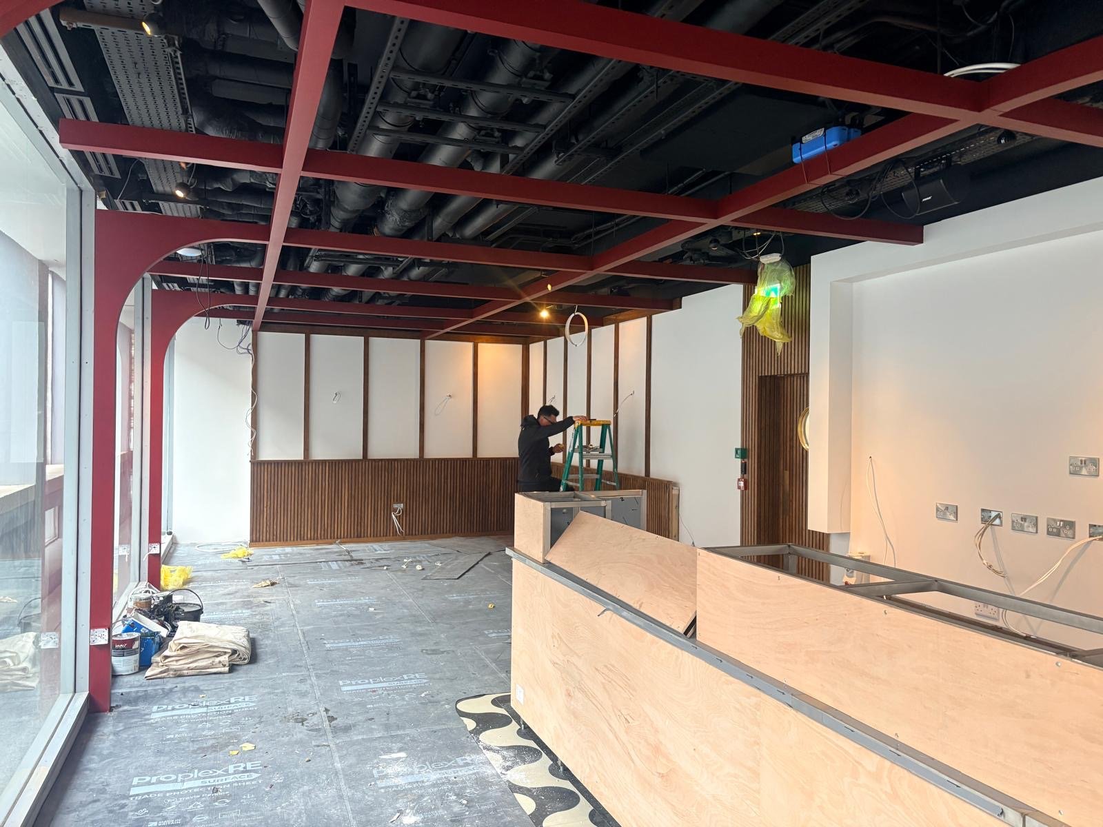 A worker stands on a ladder installing lights in a modern interior under construction, with exposed red ceiling beams, wooden wall panels, and unfinished counters—creating the inviting atmosphere of Guillam Coffee in London. A worker stands on a ladder installing lights in a modern interior under construction, with exposed red ceiling beams, wooden wall panels, and unfinished counters—creating the inviting atmosphere of Guillam Coffee in London.