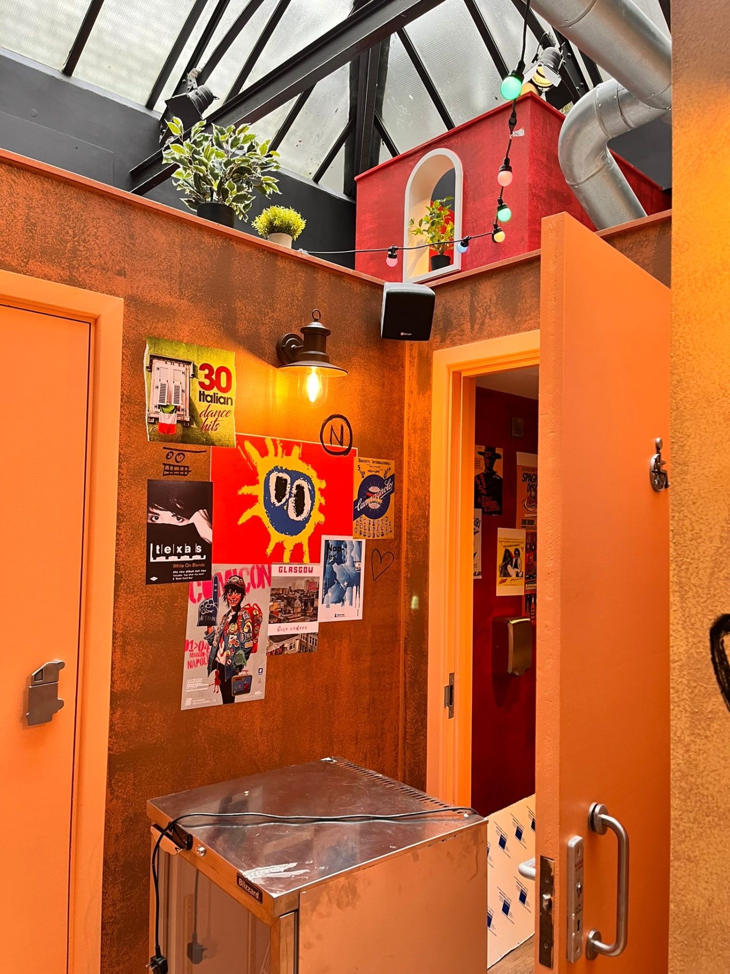 A vibrant corridor in Glasgow with orange doors, eclectic posters, a red sun artwork, a skylight, a small red house with plants above, fairy lights, and a metallic hand dryer on the wall evokes the playful spirit of Pizza Pilgrims.