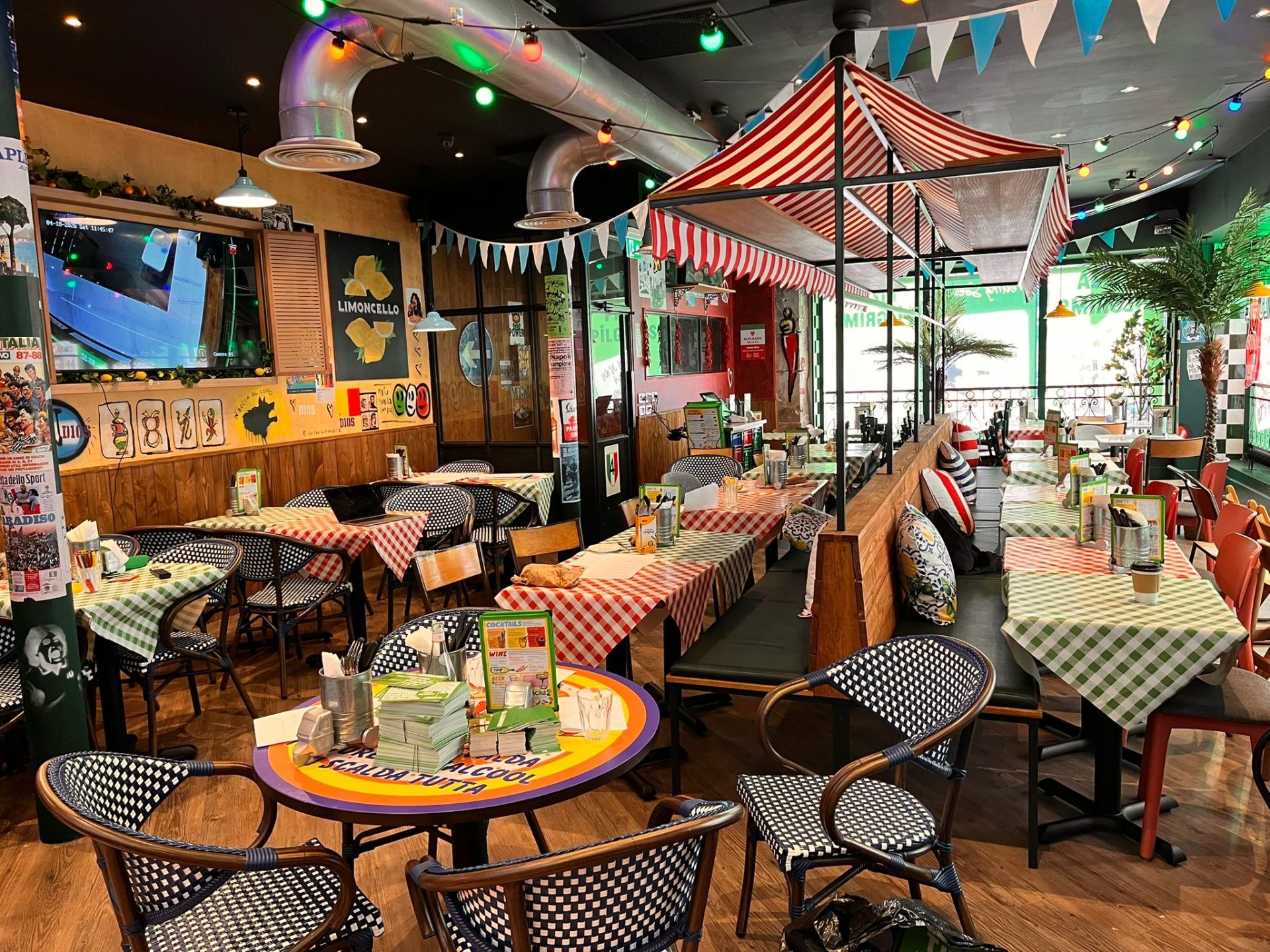 Colourful, lively restaurant with chequered tablecloths, hanging lights, bunting, and festive decor. Inspired by the energy of Glasgow, Pizza Pilgrims sets tables with menus and condiments, while picnic-style benches and chairs fill the inviting, playful space.