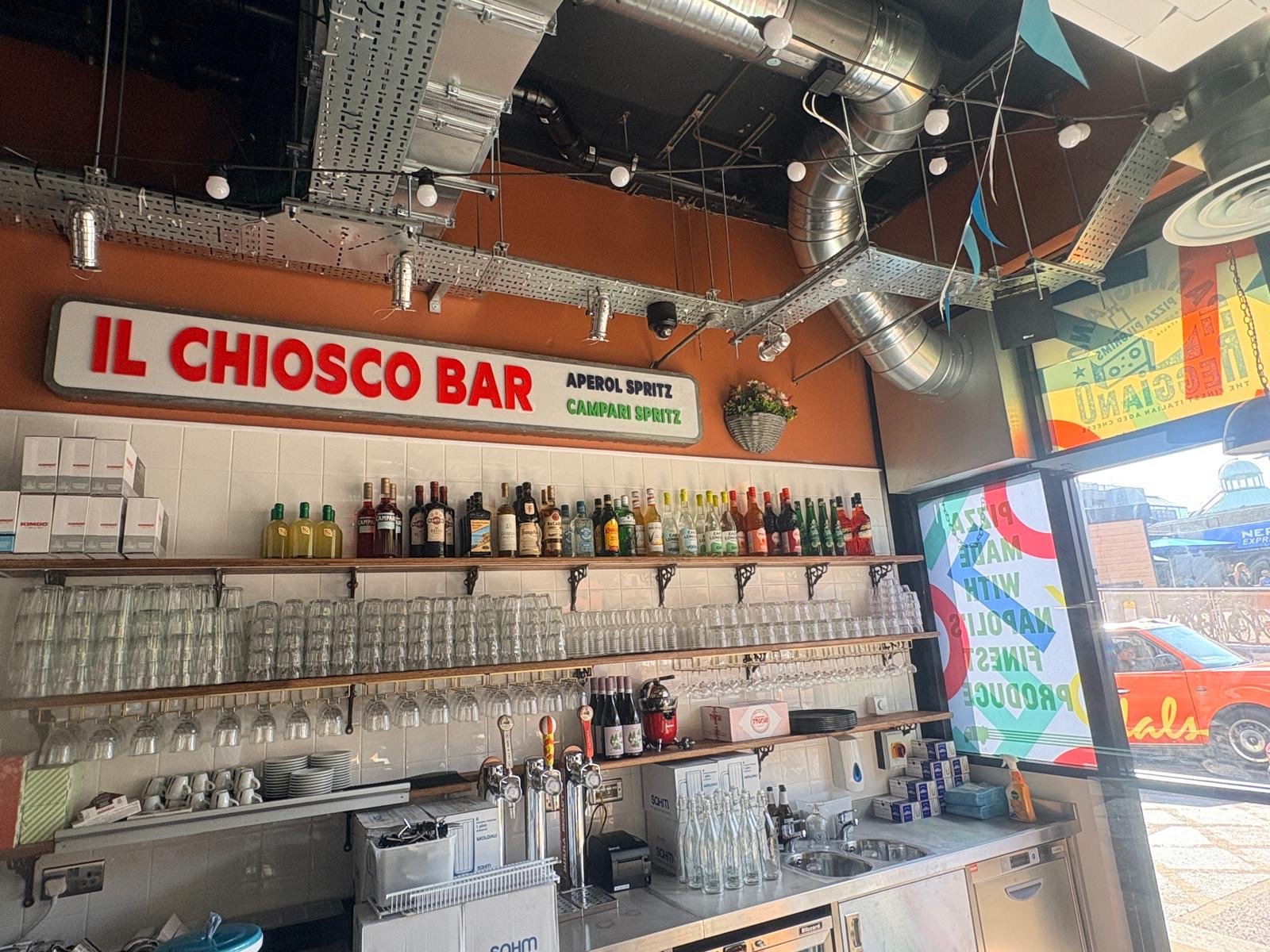 A bar with a sign reading IL CHIOSCO BAR. Bottles and glasses line the shelves, and colourful decorations hang from the ceiling. Sunlight streams through a window with a vibrant mural, creating an atmosphere that brings to mind the lively spirit of Wimbledon or the warmth found at Pizza Pilgrims.