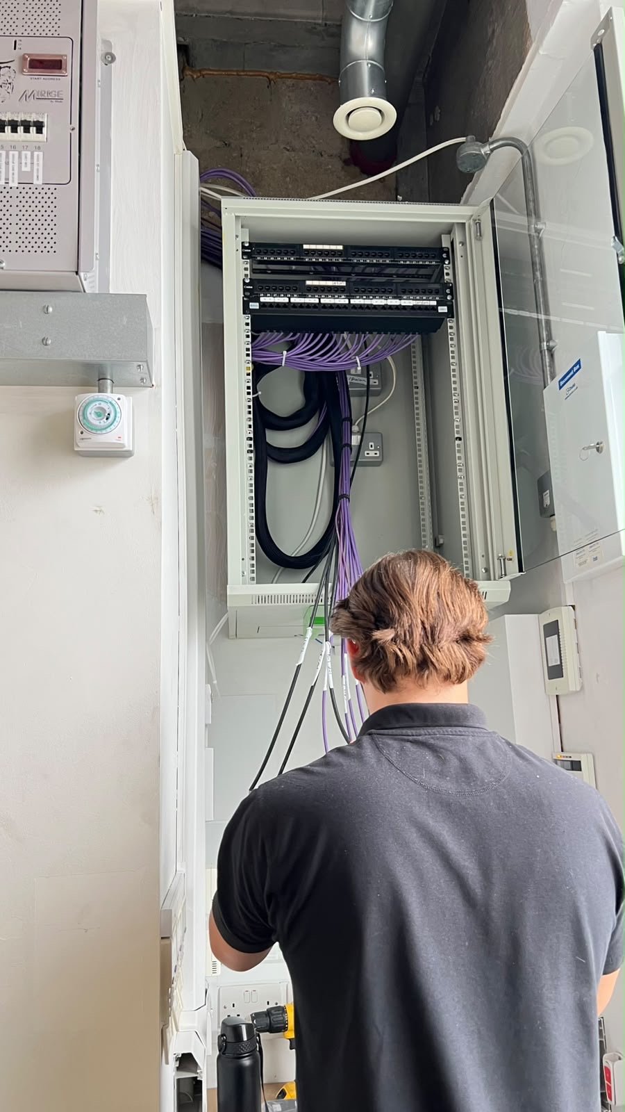 A person with brown hair works on a network server cabinet with purple cables and equipment in a utility room at Pizza Pilgrims Wimbledon. The server door is open, revealing organised wiring and components.