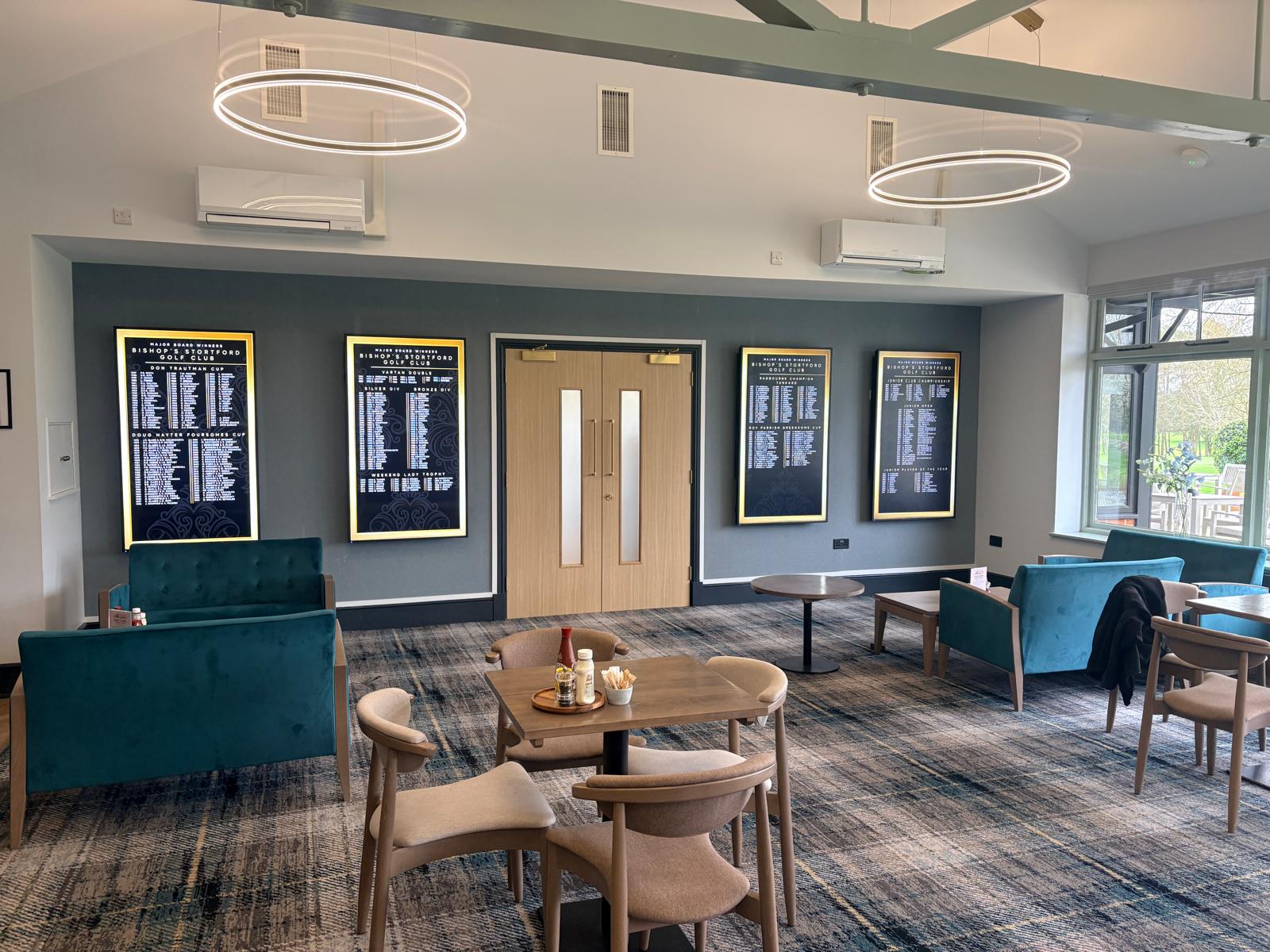 A modern lounge at Bishops Stortford Golf Club features teal chairs, wooden tables, and wall plaques. Light fittings hang from the ceiling, double doors are centred, and large windows let in natural light from the right—creating a warm and inviting space for golf enthusiasts in Hertfordshire.