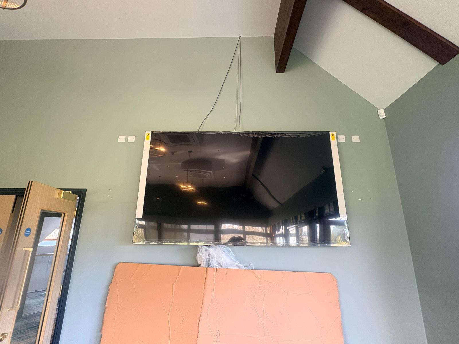 A large flat-screen TV is wall-mounted in a room with green walls and a high ceiling, reminiscent of the lush fairways at Bishops Stortford Golf Club in Hertfordshire. An orange panel leans below the TV, and a cable runs from the TV to the ceiling.