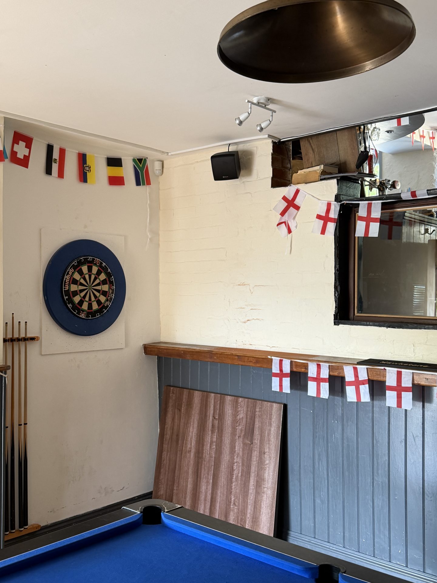 A dartboard on a wall, pool cues nearby, and a blue pool table lend an authentic pub atmosphere. International and England flags hang for decoration at the Nags Head in Lyme Regis. A wooden board leans against the wall below a shelf, whilst a mirror reflects the dartboard. A dartboard on a wall, pool cues nearby, and a blue pool table lend an authentic pub atmosphere. International and England flags hang for decoration at the Nags Head in Lyme Regis. A wooden board leans against the wall below a shelf, whilst a mirror reflects the dartboard.