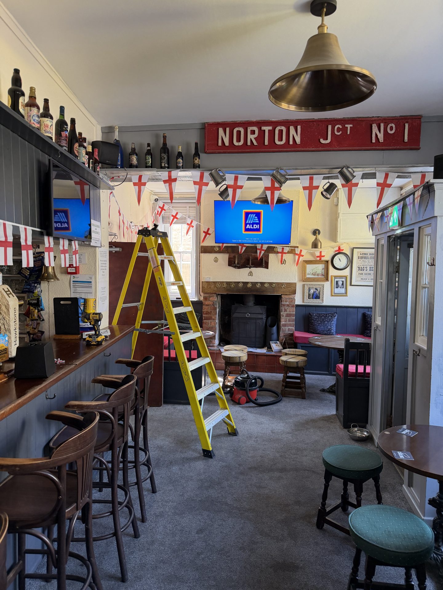 A yellow ladder stands in the centre of a decorated pub with English flags, bar stools, two green stools, and two TVs displaying the ALDI logo. Reminiscent of classic venues like the Nags Head in Lyme Regis, a red sign reads NORTON JCT NO 1 above a fireplace. A yellow ladder stands in the centre of a decorated pub with English flags, bar stools, two green stools, and two TVs displaying the ALDI logo. Reminiscent of classic venues like the Nags Head in Lyme Regis, a red sign reads NORTON JCT NO 1 above a fireplace.