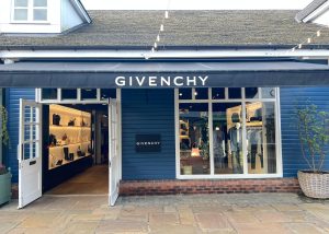 Exterior of the Givenchy boutique at Bicester Village, featuring a striking blue wooden facade, elegant black awning, open doors, and window displays showcasing stylish clothes and accessories.