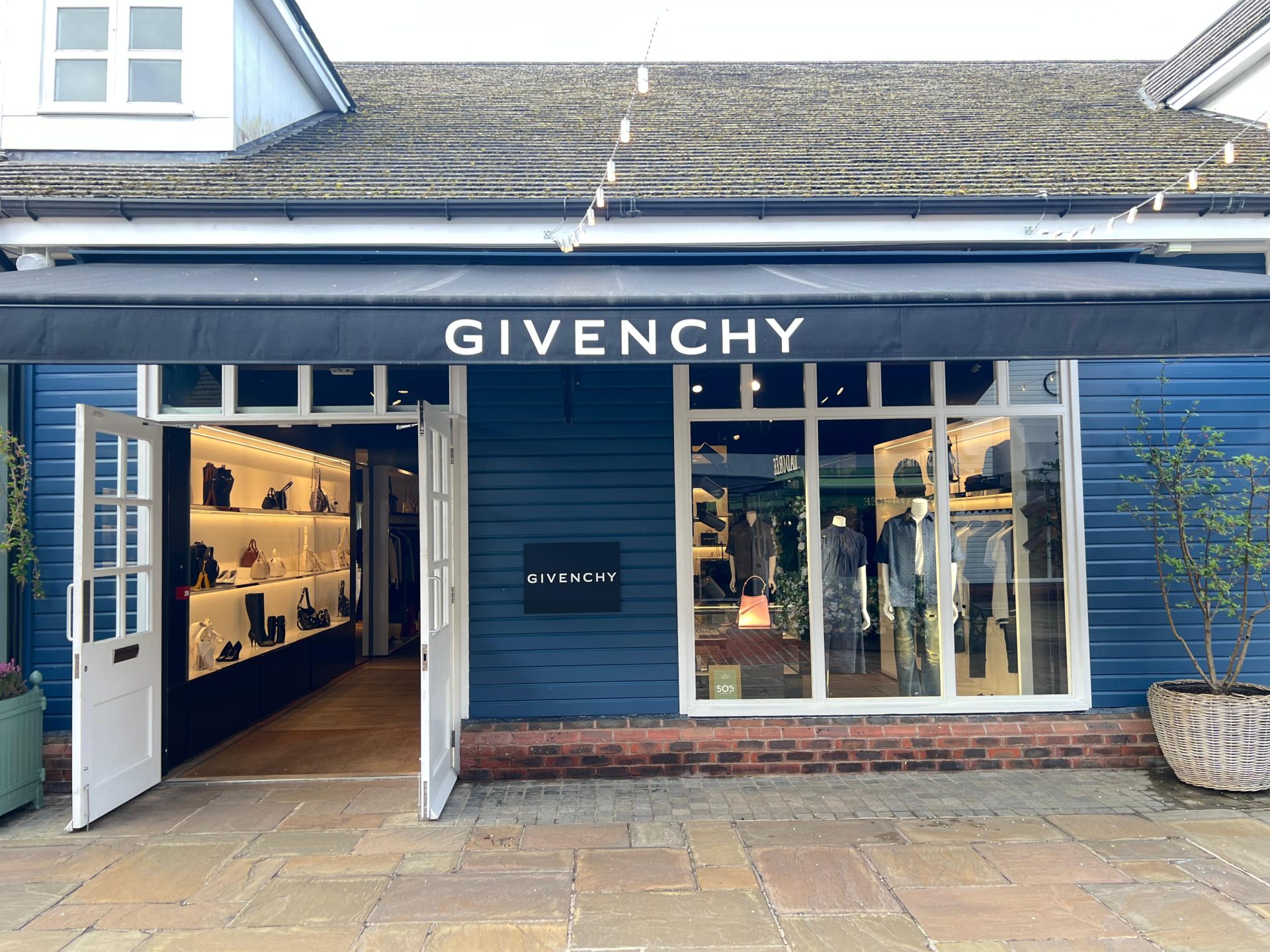 Exterior of the Givenchy boutique at Bicester Village, featuring a striking blue wooden facade, elegant black awning, open doors, and window displays showcasing stylish clothes and accessories. Exterior of the Givenchy boutique at Bicester Village, featuring a striking blue wooden facade, elegant black awning, open doors, and window displays showcasing stylish clothes and accessories.