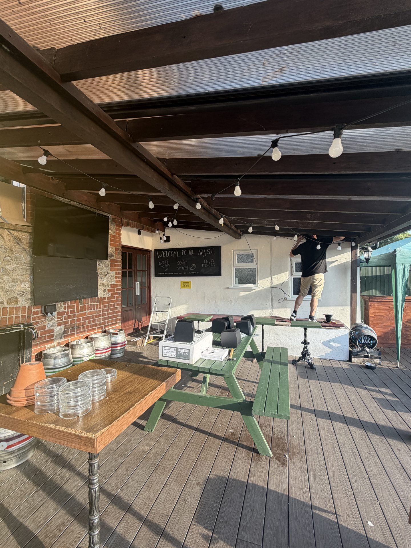A person stands on a table adjusting fairy lights under a pergola on a wooden patio at the Nags Head pub. Nearby are green picnic tables, beer barrels, clear glasses, and a blackboard on the wall—creating a welcoming scene reminiscent of coastal gatherings in Lyme Regis. A person stands on a table adjusting fairy lights under a pergola on a wooden patio at the Nags Head pub. Nearby are green picnic tables, beer barrels, clear glasses, and a blackboard on the wall—creating a welcoming scene reminiscent of coastal gatherings in Lyme Regis.