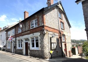 A two-storey stone and brick pub called Nags Head Inn stands on a sunny street corner in Lyme Regis, with a flag, lanterns, and distant hills visible in the background.