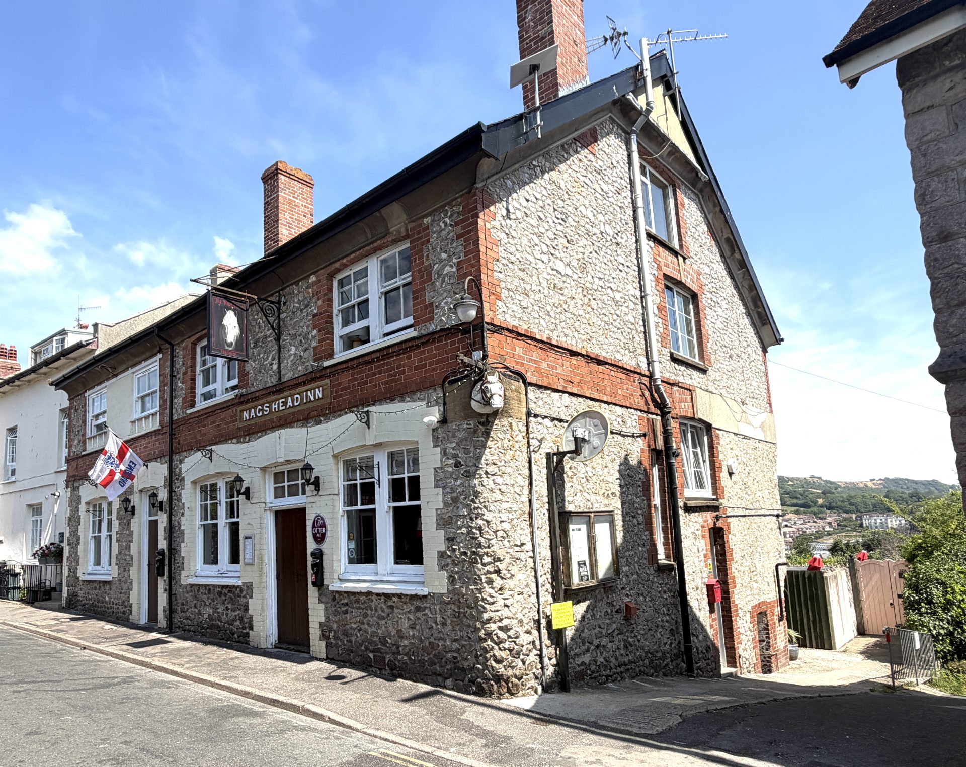A two-storey stone and brick pub called Nags Head Inn stands on a sunny street corner in Lyme Regis, with a flag, lanterns, and distant hills visible in the background. A two-storey stone and brick pub called Nags Head Inn stands on a sunny street corner in Lyme Regis, with a flag, lanterns, and distant hills visible in the background.