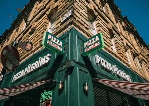A corner view of a Pizza Pilgrims restaurant in Glasgow, with green signage and a tan stone building under a clear blue sky. A no entry road sign and striped awning are also visible.