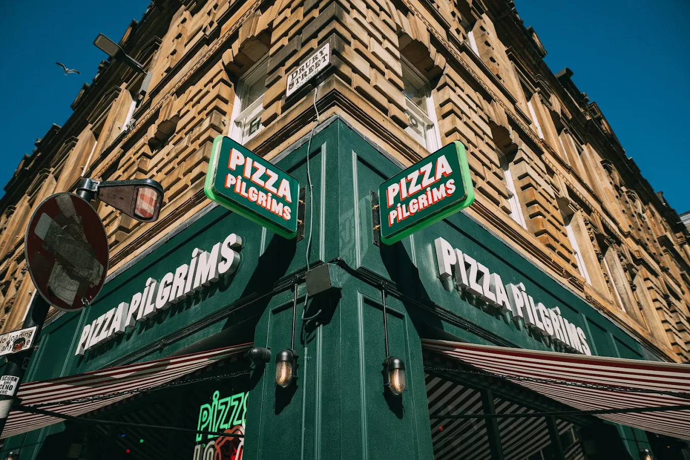 A corner view of a Pizza Pilgrims restaurant in Glasgow, with green signage and a tan stone building under a clear blue sky. A no entry road sign and striped awning are also visible.