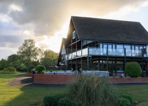 Modern building with large glass windows and a steep roof, surrounded by green grass and trees, with outdoor seating bathed in sunlight under a partly cloudy sky—an inviting setting that perfectly complements the tranquil landscape of Hertfordshire near Bishops Stortford Golf Club.