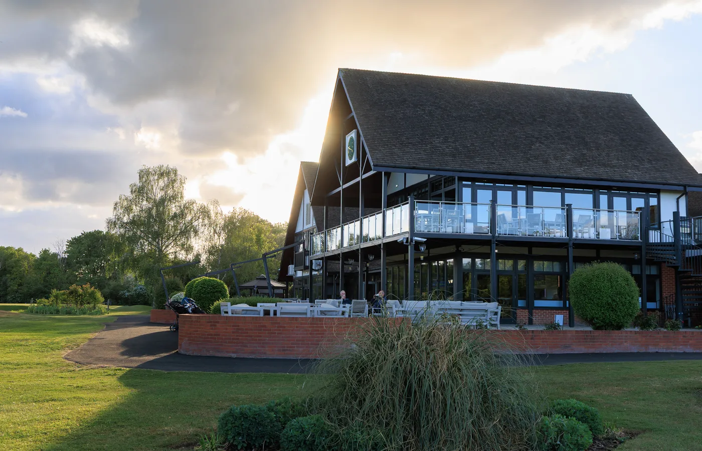Modern building with large glass windows and a steep roof, surrounded by green grass and trees, with outdoor seating bathed in sunlight under a partly cloudy sky—an inviting setting that perfectly complements the tranquil landscape of Hertfordshire near Bishops Stortford Golf Club.
