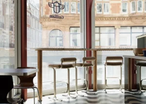 Modern café interior with tall windows, patterned floor, high tables, and chairs. Sunlight streams in, and outside, brick buildings are visible—evoking the vibrant charm of London. The Guillam Coffee logo is on the window.