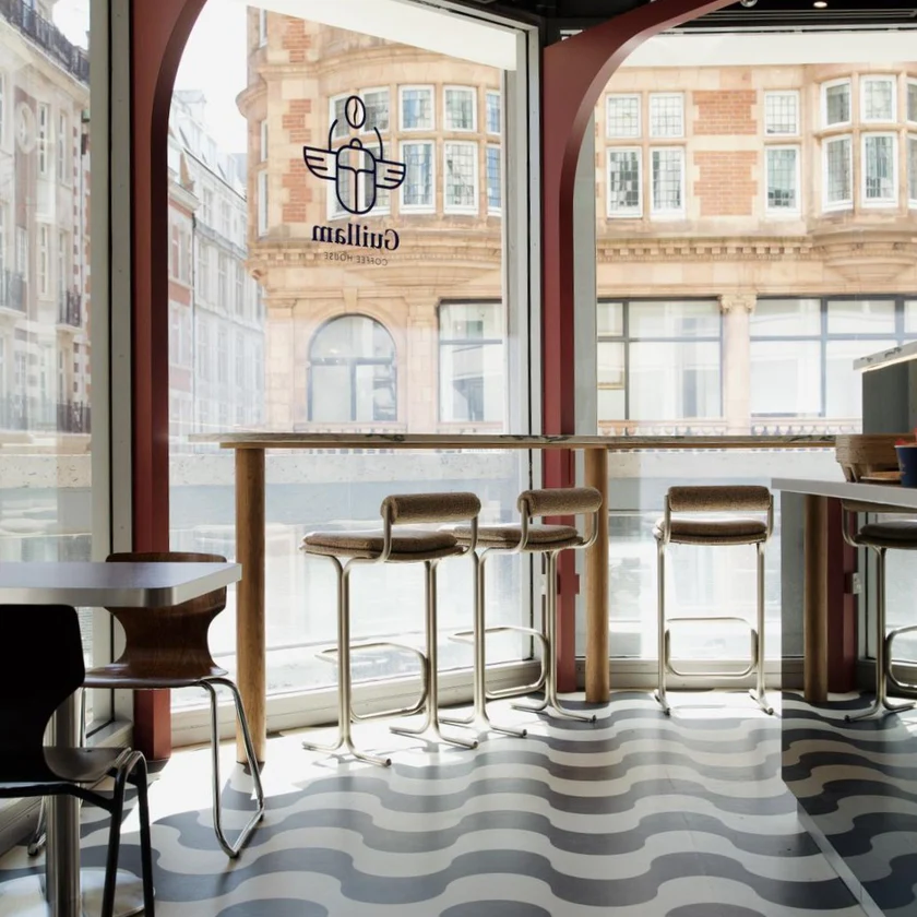 Modern café interior with tall windows, patterned floor, high tables, and chairs. Sunlight streams in, and outside, brick buildings are visible—evoking the vibrant charm of London. The Guillam Coffee logo is on the window. Modern café interior with tall windows, patterned floor, high tables, and chairs. Sunlight streams in, and outside, brick buildings are visible—evoking the vibrant charm of London. The Guillam Coffee logo is on the window.