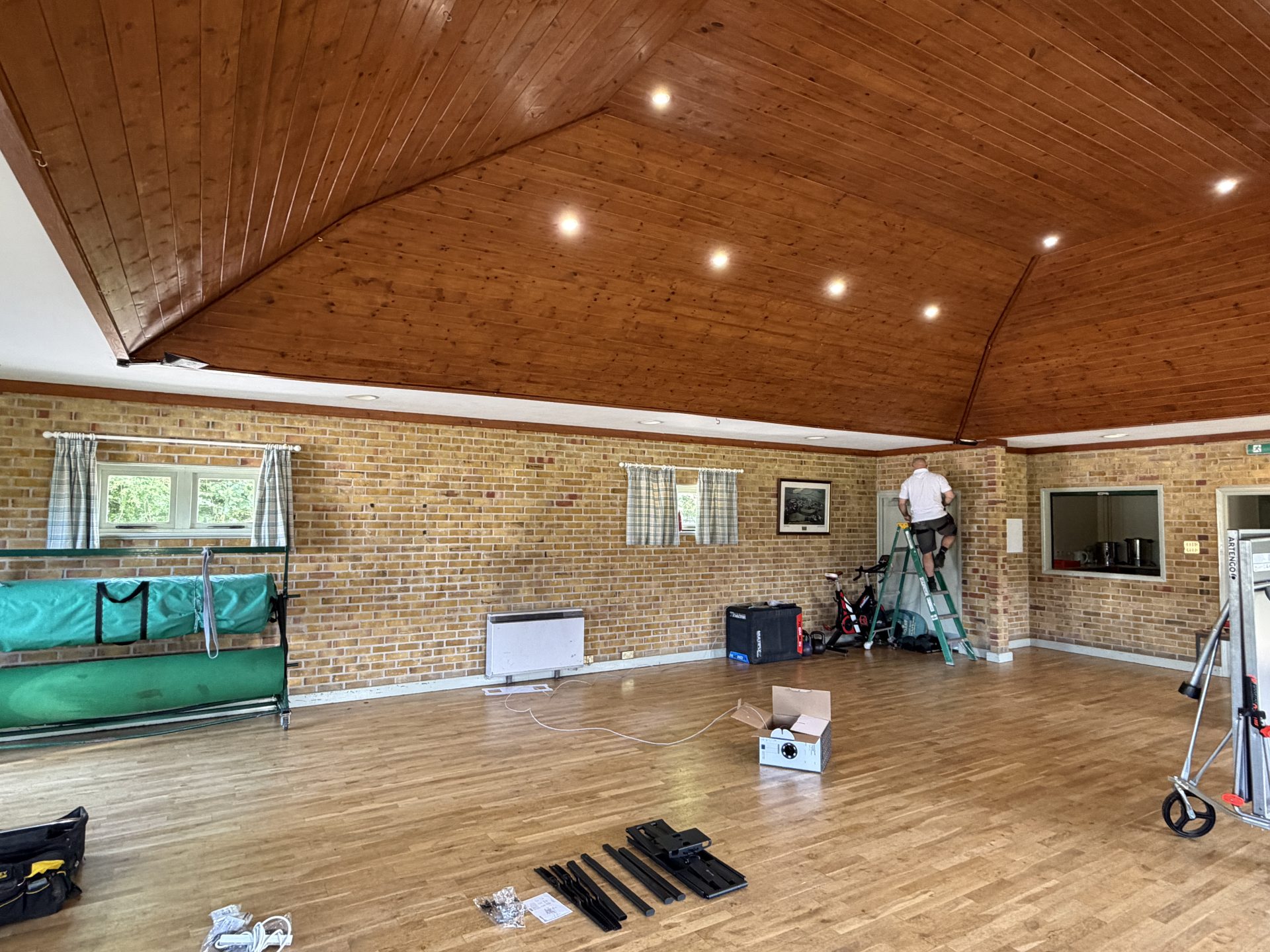 A spacious hall with wooden floors and ceiling, reminiscent of the charming Pleshey Village Hall in Essex. A person on a ladder works near a brick wall, while tools and equipment are scattered on the floor. There are large windows with curtains and a green settee on the left.