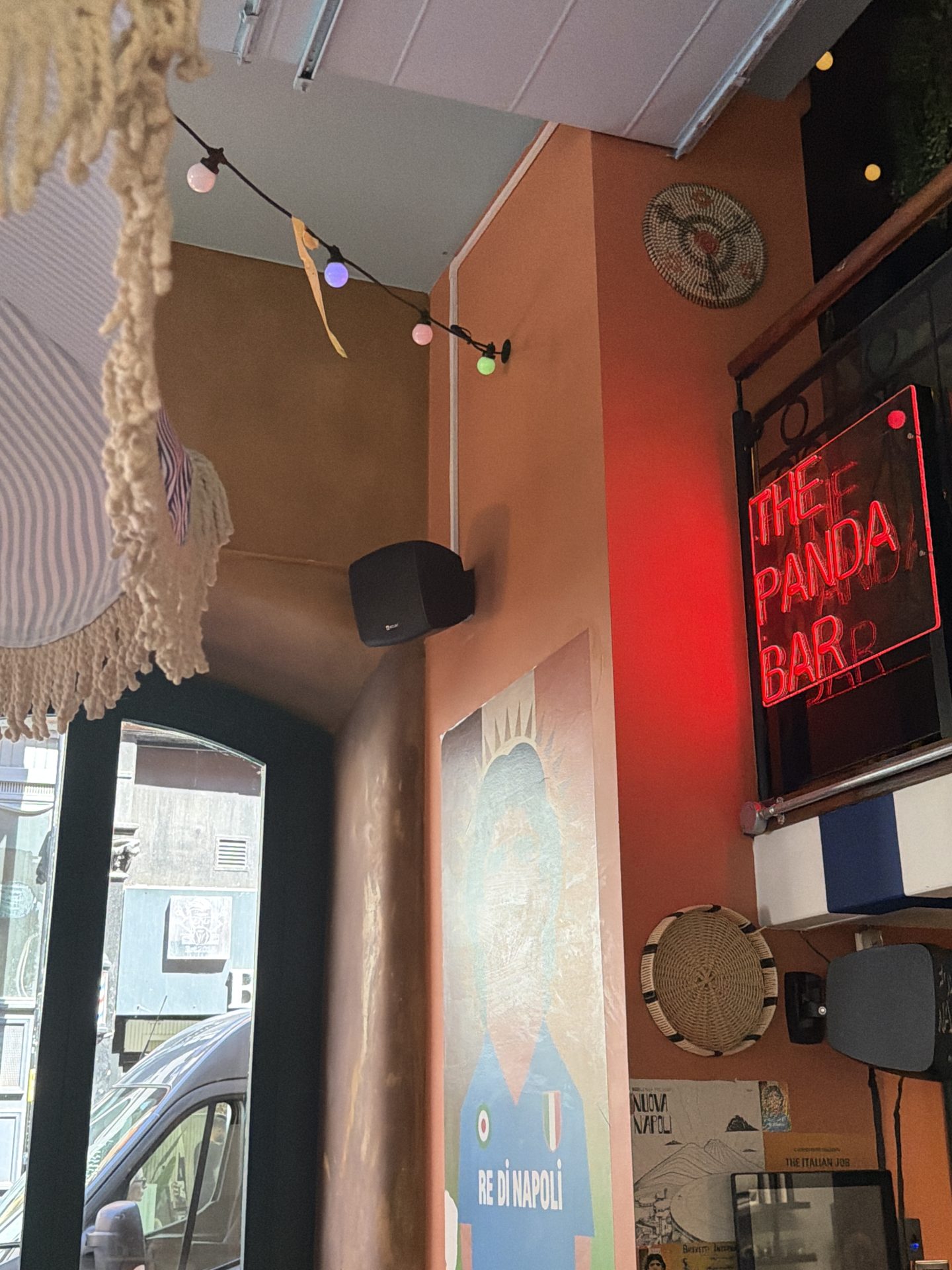 Inside a bar with orange walls near Deansgate, a neon sign reads THE PANDA BAR. There are decorative baskets on the wall, fairy lights, a speaker, and a partial mural of a person in a blue shirt. Through the window, you can spot a van parked outside in the bustling heart of Manchester. Inside a bar with orange walls near Deansgate, a neon sign reads THE PANDA BAR. There are decorative baskets on the wall, fairy lights, a speaker, and a partial mural of a person in a blue shirt. Through the window, you can spot a van parked outside in the bustling heart of Manchester.