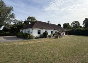 A single-storey white building with a brown roof stands near a lawn, surrounded by trees and shrubs, under a partly cloudy sky. A wooden picnic table sits on the grass outside, capturing the welcoming atmosphere of Essex’s Pleshey Village Hall.