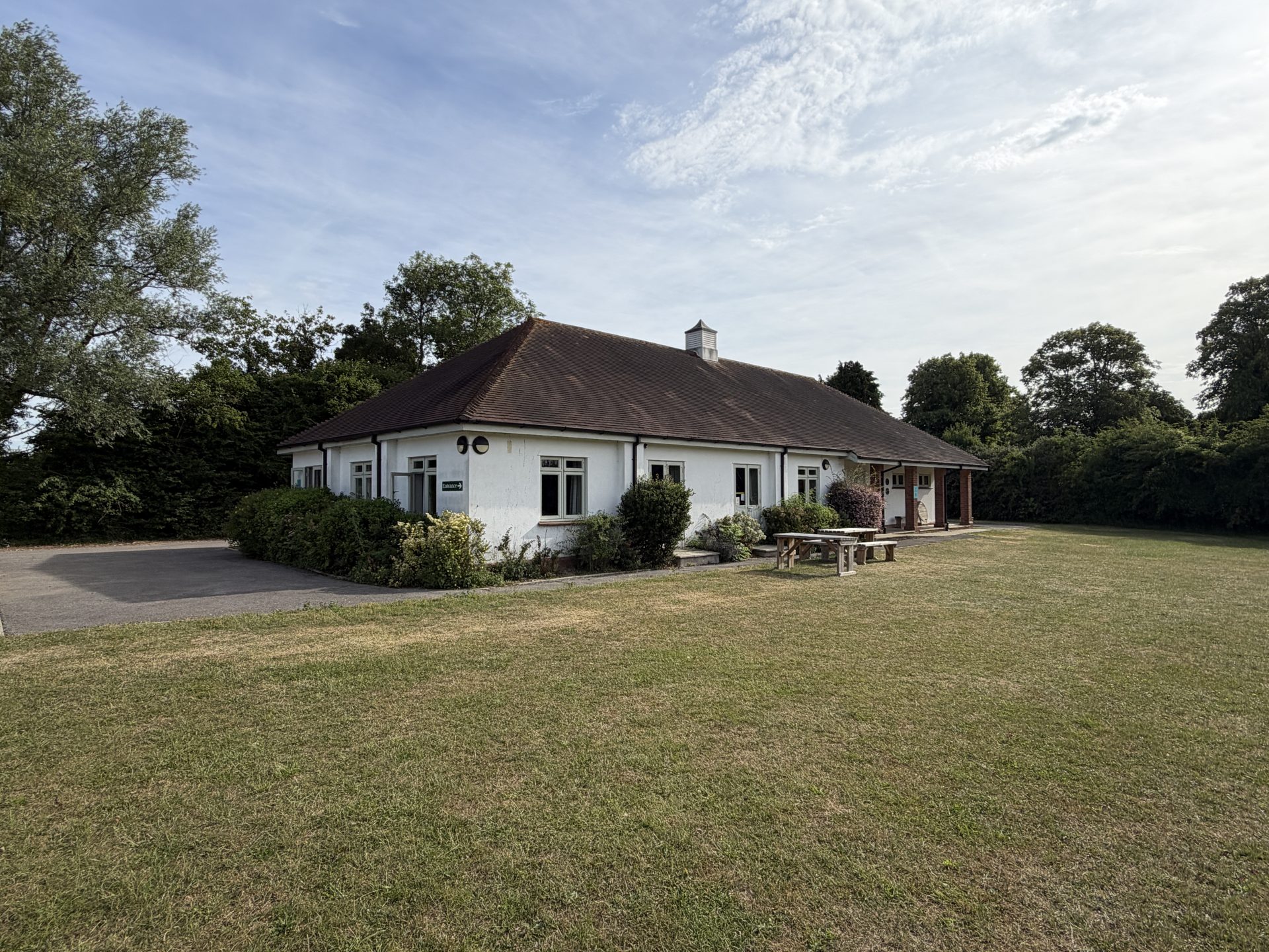 A single-storey white building with a brown roof stands near a lawn, surrounded by trees and shrubs, under a partly cloudy sky. A wooden picnic table sits on the grass outside, capturing the welcoming atmosphere of Essex’s Pleshey Village Hall.