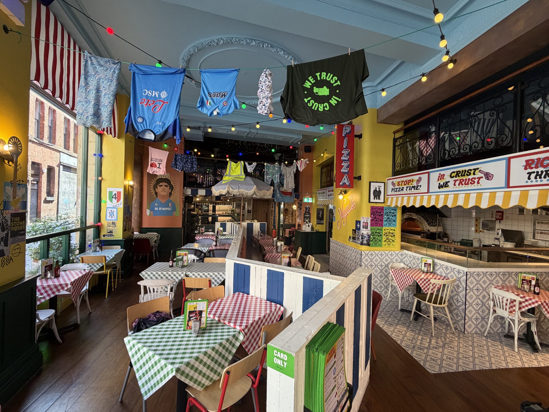 A vibrant pizzeria on Deansgate, Manchester, with chequered tablecloths, colourful decor, hanging laundry, fairy lights, and pizza-themed signs. The interior at Pizza Pilgrims is lively, quirky, and inviting with various posters on the walls. A vibrant pizzeria on Deansgate, Manchester, with chequered tablecloths, colourful decor, hanging laundry, fairy lights, and pizza-themed signs. The interior at Pizza Pilgrims is lively, quirky, and inviting with various posters on the walls.
