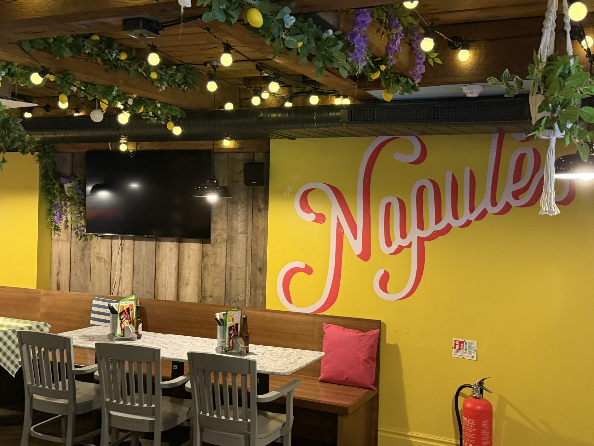 A cosy restaurant interior on Deansgate in Manchester, featuring a yellow wall reading Napule, wooden table, grey chairs, hanging greenery, fairy lights, a television, menus, a fire extinguisher, and decorative cushions—perfect for enjoying the vibrant atmosphere at Pizza Pilgrims. A cosy restaurant interior on Deansgate in Manchester, featuring a yellow wall reading Napule, wooden table, grey chairs, hanging greenery, fairy lights, a television, menus, a fire extinguisher, and decorative cushions—perfect for enjoying the vibrant atmosphere at Pizza Pilgrims.