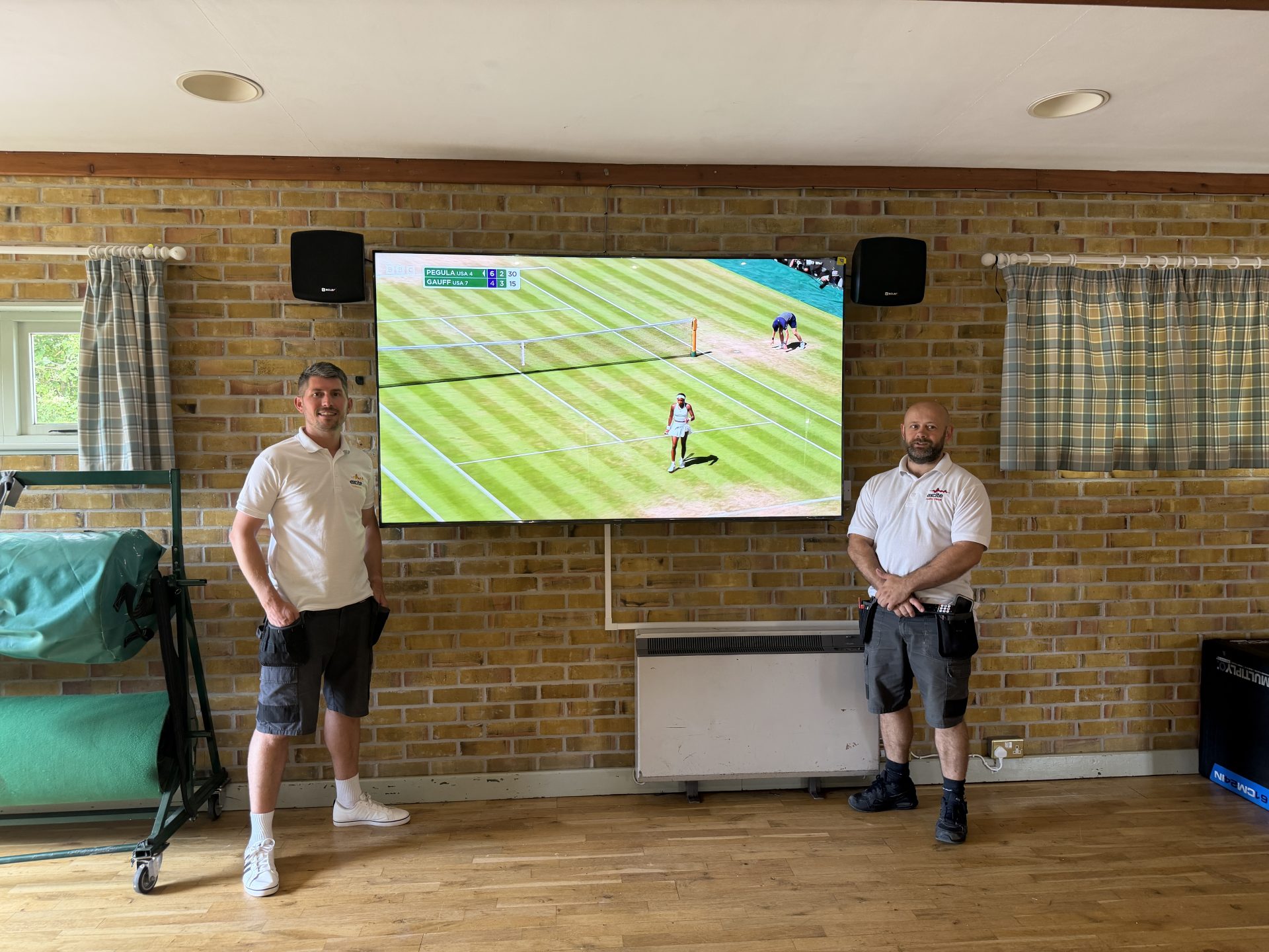 Two men in white shirts and shorts stand beside a wall-mounted TV displaying a tennis match, in a room with brick walls, wooden floor, curtains, and speakers at Pleshey Village Hall in Essex.