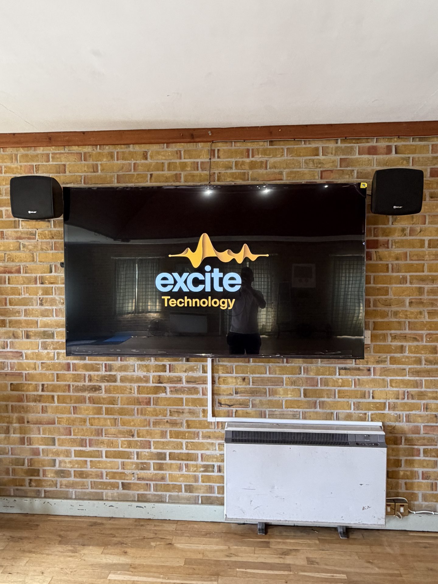 A flat-screen TV mounted on a brick wall at Pleshey Village Hall in Essex displays the excite Technology logo, with two black speakers on either side and a heater unit below. A person’s reflection is visible on the TV screen.