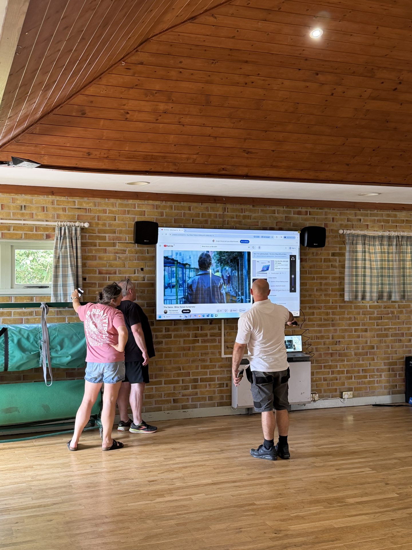 Three people stand in a room with wooden floors and a brick wall at Pleshey Village Hall in Essex, watching a large screen showing a video. Two of them point at the screen; sunlight enters through small windows with striped curtains.