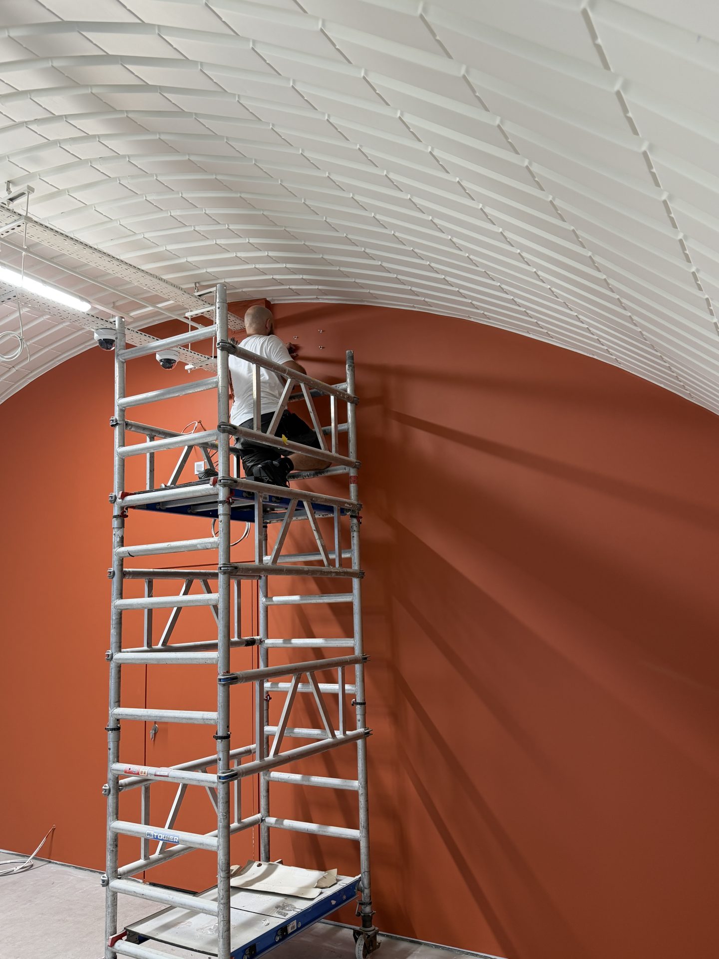 A person on a scaffold is working on a high, curved ceiling with white tiles and a red-orange wall below in The Solt Room, Hackney, London.