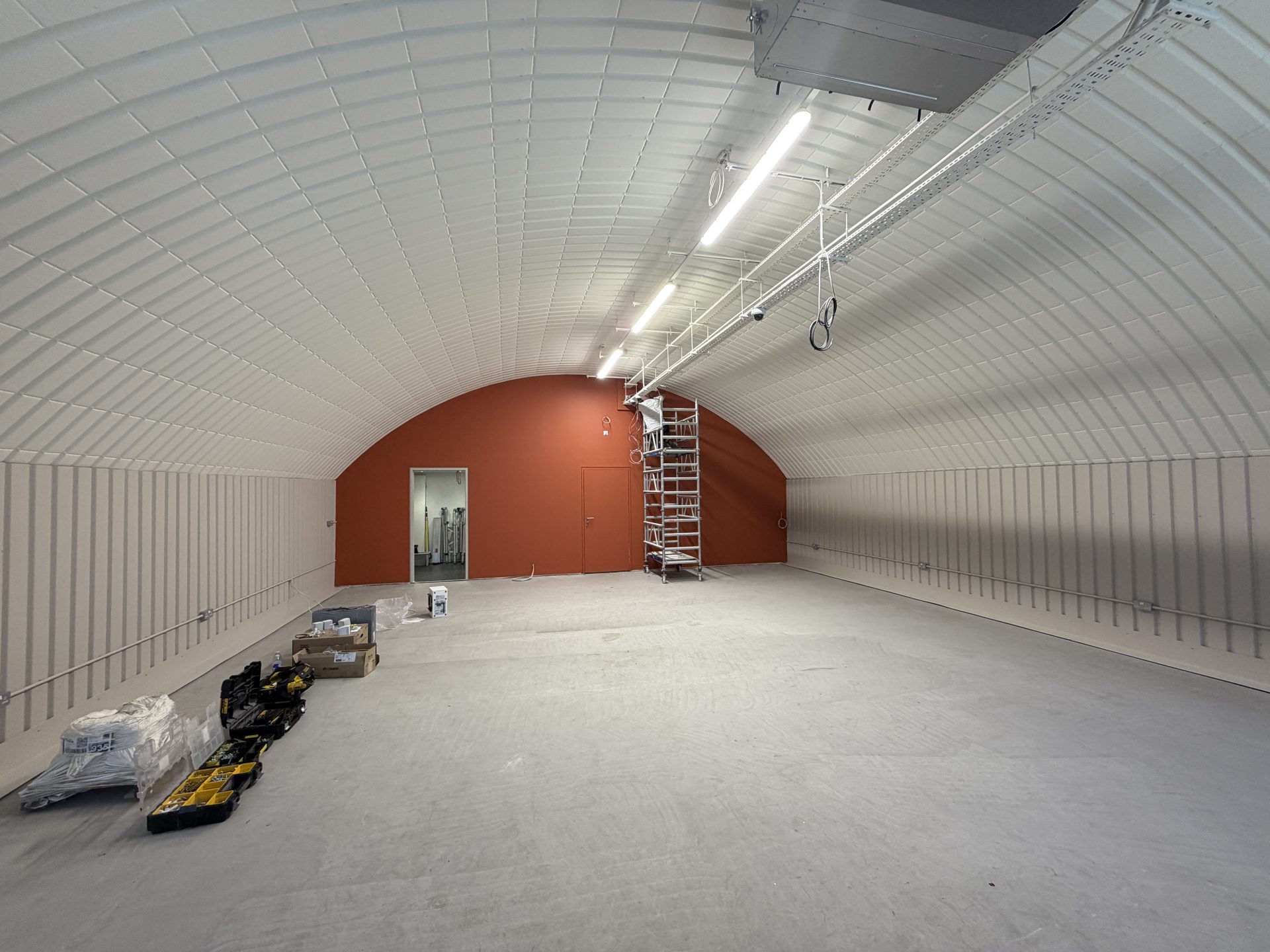 A large, empty, arched room—known as the Solt Room—in Hackney, London, features a striking red back wall, a metal scaffold, tools, and boxes scattered across the floor. Overhead fluorescent lights illuminate the space.
