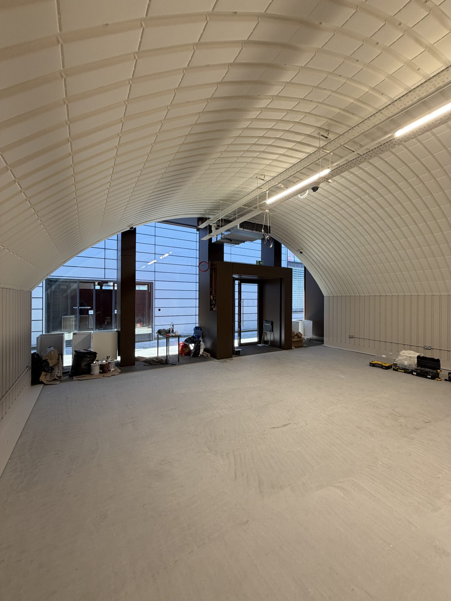 A spacious, empty Solt Room in Hackney, London, featuring a curved ceiling and large front windows with scattered construction materials on the sides. Natural light pours in through the glass doors and windows at the far end.
