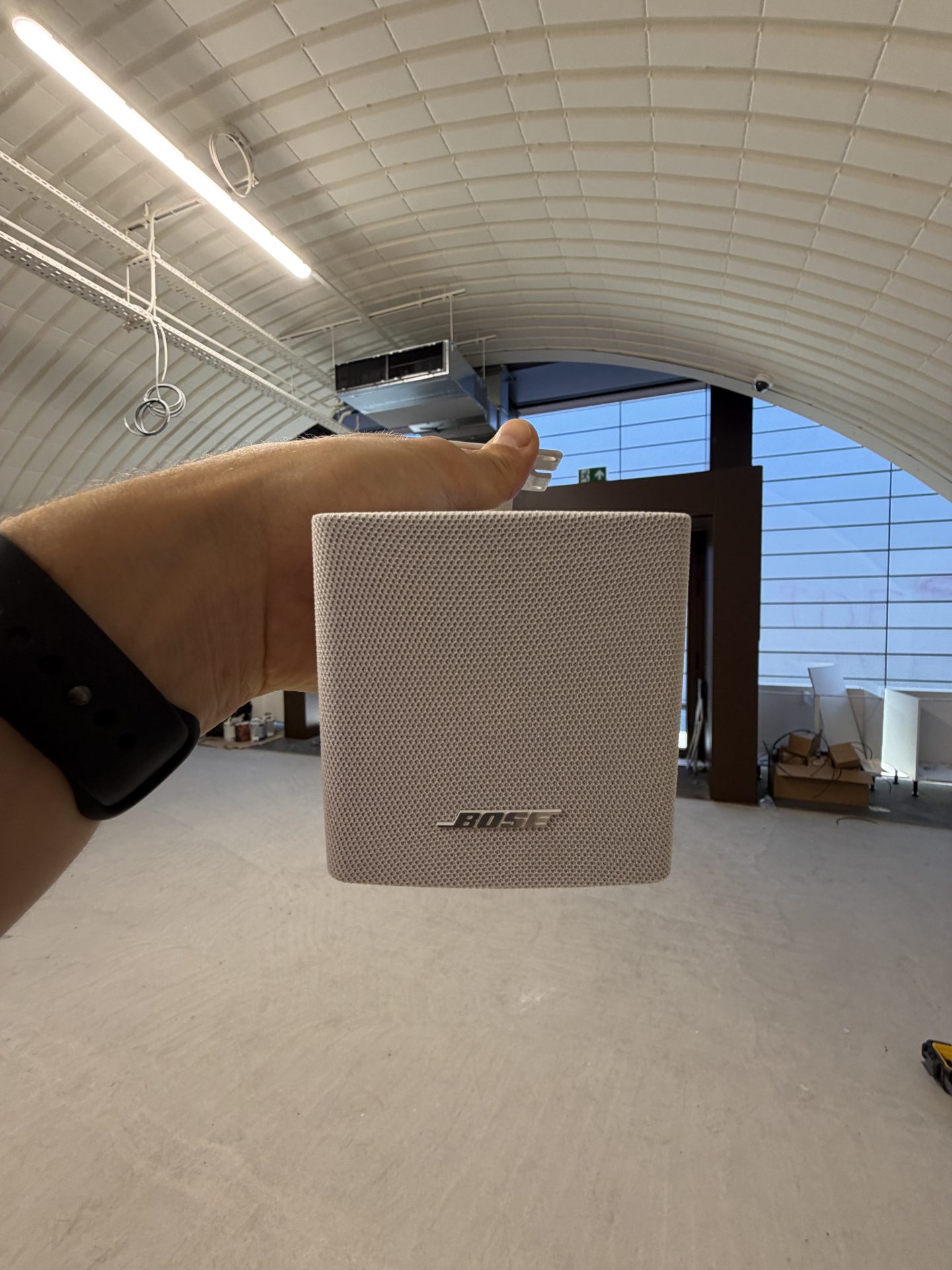 A hand holding a white Bose speaker inside the large, modern, mostly empty Solt Room in Hackney, London, with a curved ceiling and some boxes and equipment visible in the background.