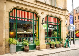 Street view of Pizza Pilgrims in Manchester, located on Deansgate, featuring large windows, red and white striped awnings, green trim, and wooden barrels with plants outside. The urban setting includes surrounding buildings and a bustling pavement.