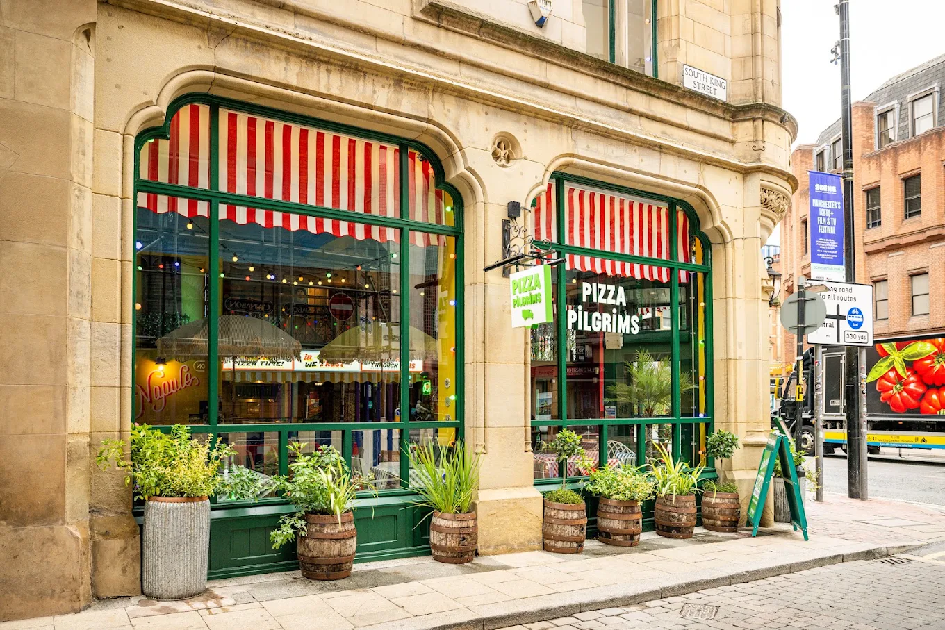 Street view of Pizza Pilgrims in Manchester, located on Deansgate, featuring large windows, red and white striped awnings, green trim, and wooden barrels with plants outside. The urban setting includes surrounding buildings and a bustling pavement. Street view of Pizza Pilgrims in Manchester, located on Deansgate, featuring large windows, red and white striped awnings, green trim, and wooden barrels with plants outside. The urban setting includes surrounding buildings and a bustling pavement.