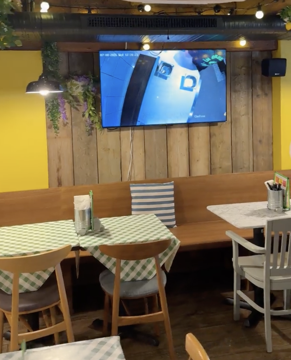 A cosy restaurant on Deansgate with chequered tablecloths, wooden benches, and a TV mounted on a wooden wall displaying security footage. The décor includes hanging lights, plants, and yellow walls—making Pizza Pilgrims in Manchester the perfect spot to relax and enjoy delicious food. A cosy restaurant on Deansgate with chequered tablecloths, wooden benches, and a TV mounted on a wooden wall displaying security footage. The décor includes hanging lights, plants, and yellow walls—making Pizza Pilgrims in Manchester the perfect spot to relax and enjoy delicious food.