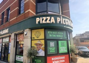 Exterior of a Pizza Pilgrims restaurant on a sunny day in Wimbledon, featuring large windows, colourful signs, and a photo of a smiling man in a hat on the shopfront. Signs read 6th Best Pizza in the World and Buongiorno Wimbledon.