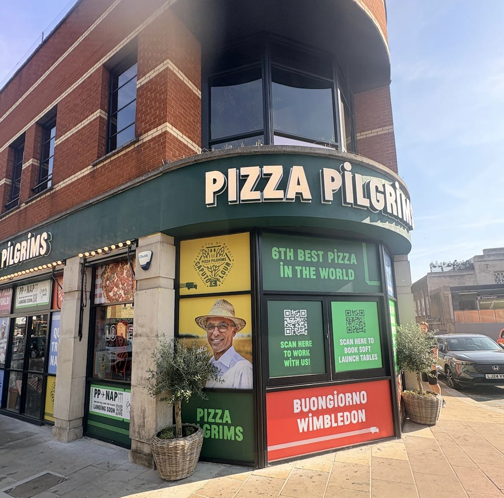 Exterior of a Pizza Pilgrims restaurant on a sunny day in Wimbledon, featuring large windows, colourful signs, and a photo of a smiling man in a hat on the shopfront. Signs read 6th Best Pizza in the World and Buongiorno Wimbledon.