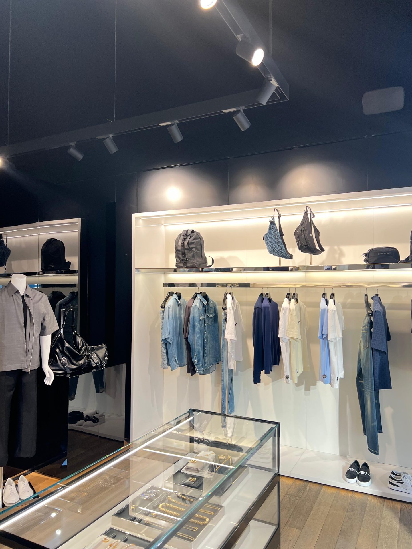 Boutique clothing shop with shirts, jackets, and trousers displayed on rails, bags on a shelf above, shoes below, and a glass cabinet with accessories in the foreground. Mannequin on the left wears a grey shirt and black trousers—a stylish destination reminiscent of Bicester Village’s fashion-forward atmosphere. Boutique clothing shop with shirts, jackets, and trousers displayed on rails, bags on a shelf above, shoes below, and a glass cabinet with accessories in the foreground. Mannequin on the left wears a grey shirt and black trousers—a stylish destination reminiscent of Bicester Village’s fashion-forward atmosphere.