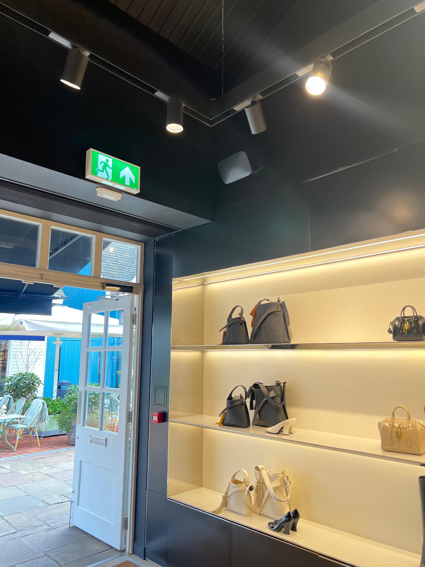 A shelf displays Givenchy handbags and shoes inside a shop with dark walls; an open door leads outside where patio chairs and tables are visible in Bicester Village. An illuminated green exit sign hangs above the door. A shelf displays Givenchy handbags and shoes inside a shop with dark walls; an open door leads outside where patio chairs and tables are visible in Bicester Village. An illuminated green exit sign hangs above the door.