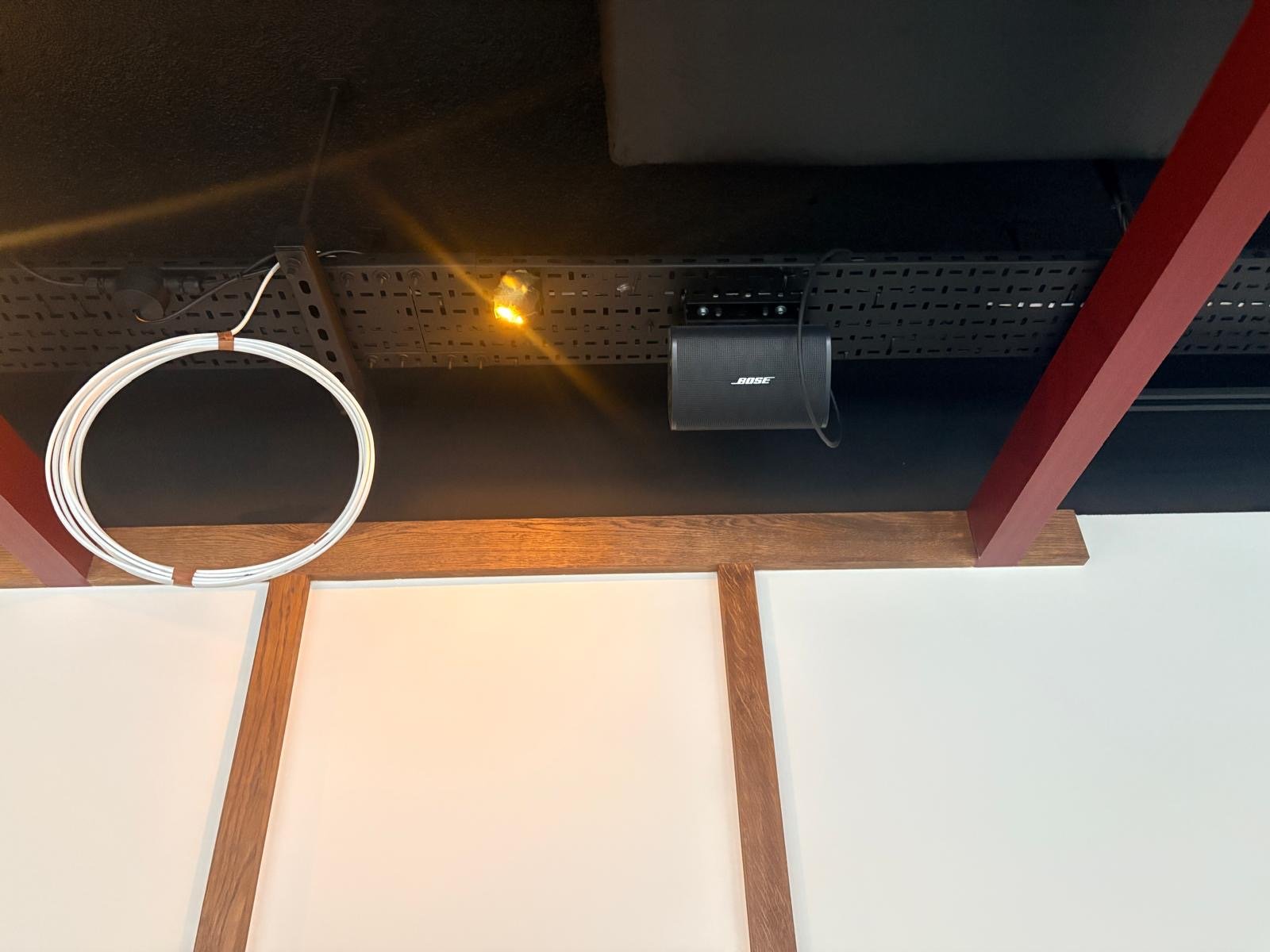 Ceiling view with a white cable loop, a yellow light, a black Bose speaker, and red and wooden beams against a white wall—capturing the cosy ambience reminiscent of Guillam Coffee in London. Ceiling view with a white cable loop, a yellow light, a black Bose speaker, and red and wooden beams against a white wall—capturing the cosy ambience reminiscent of Guillam Coffee in London.