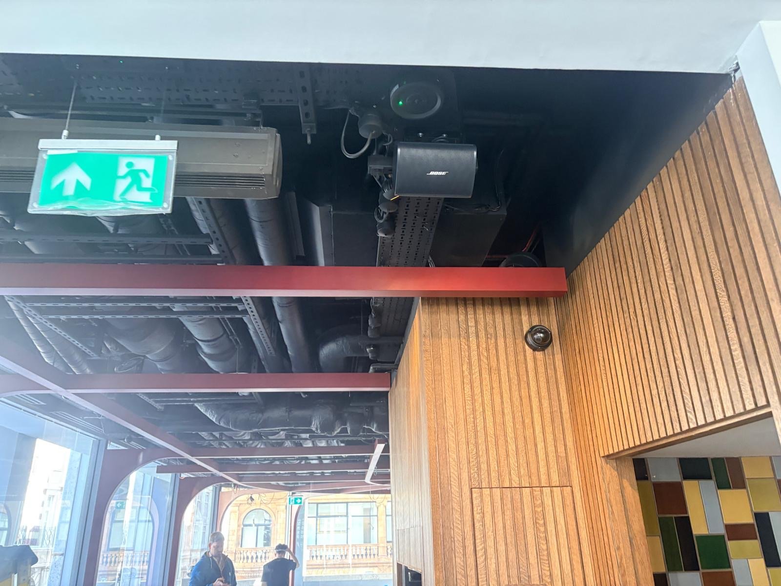 Ceiling with exposed black pipes, a green emergency exit sign, a Bose speaker, and a CCTV camera. Below, the wooden panelled wall and colourful tiles create a warm atmosphere at Guillam Coffee in London, with large windows letting in natural light—the perfect spot to enjoy your favourite coffee. Ceiling with exposed black pipes, a green emergency exit sign, a Bose speaker, and a CCTV camera. Below, the wooden panelled wall and colourful tiles create a warm atmosphere at Guillam Coffee in London, with large windows letting in natural light—the perfect spot to enjoy your favourite coffee.