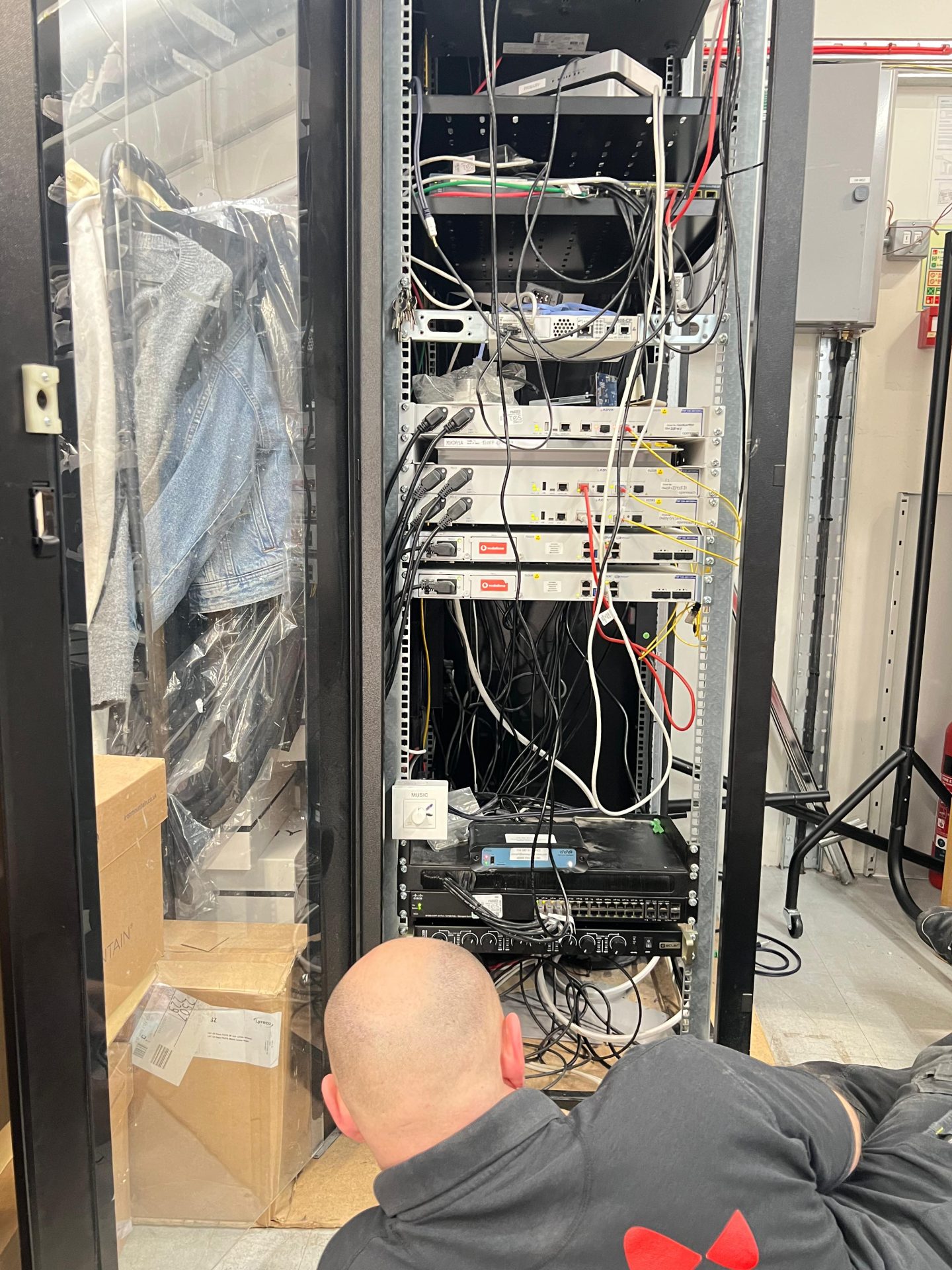 A person with a shaven head lies on the floor, working on cables in an open server rack next to a glass wardrobe with hanging clothes and cardboard boxes, reminiscent of the backroom organisation found in a sophisticated boutique at Bicester Village. A person with a shaven head lies on the floor, working on cables in an open server rack next to a glass wardrobe with hanging clothes and cardboard boxes, reminiscent of the backroom organisation found in a sophisticated boutique at Bicester Village.